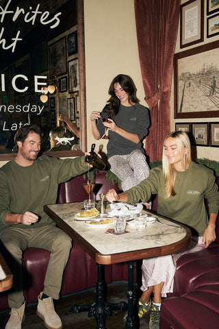 Three people in matching Cocktails & Caviar Crewneck Sweatshirts enjoy drinks and dessert at a cozy cafe. One woman stands taking a photo while the others sit, smiling and pouring drinks amid framed art and a warm atmosphere.