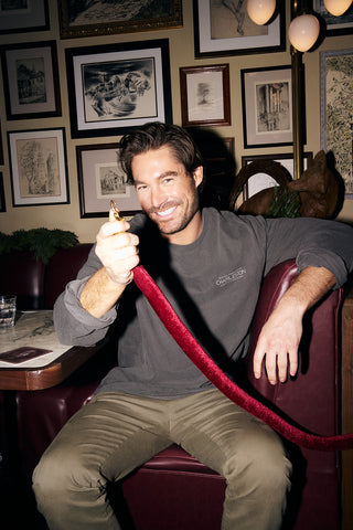 A man with brown hair and a beard smiles on a maroon booth, wearing the Cocktails & Caviar Long Sleeve Tee. Framed art covers the wall behind him, and a nearby table holds a glass of water and a menu.