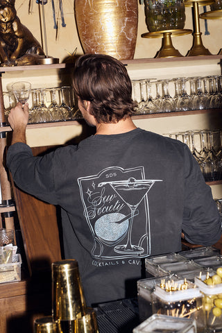 A brown-haired man, seen from behind, chooses a glass behind the bar. He sports the Cocktails & Caviar Long Sleeve Tee, featuring a martini glass and “Sun Society Cocktails & Caviar”—your ideal holiday party shirt.