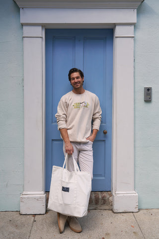 A man wearing a Holiday Rainbow Row Crewneck Sweatshirt, white pants, and boots stands smiling with a large white tote bag in front of a blue door framed by light-colored walls.