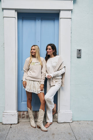 Two women pose by a light blue door. One wears the Holiday Rainbow Row Crewneck Sweatshirt with a lace skirt and cowboy boots; the other has a sweater over her shoulders and cream pants. Both are smiling in neutral-colored outfits.