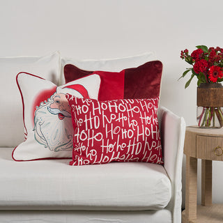 Three decorative pillows with red, white, and holiday-themed designs, including Santa and Ho Ho Ho text, sit on a white sofa next to a wooden side table with a vase of red flowers.