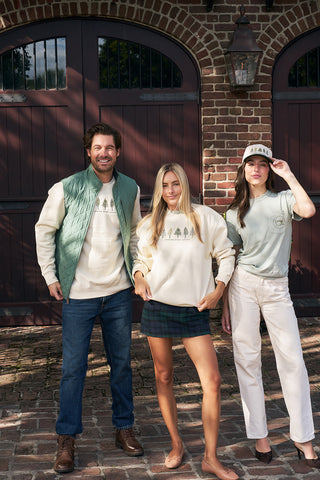 Three people smile in front of dark wooden doors and a brick wall, dressed in cozy fall outfits. One wears the Christmas Tree Farm Embroidered Crewneck, while the others sport a vest, jeans, plaid skirt, and a hat on a cobblestone path.