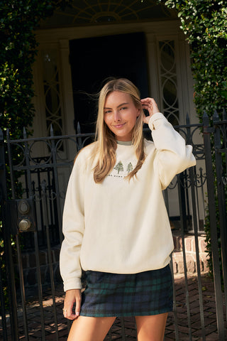 A woman stands by a black iron gate and greenery, wearing the Christmas Tree Farm Embroidered Crewneck with a tree design and a blue-green plaid skirt. She touches her hair and smiles slightly at the camera.
