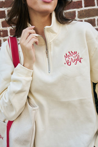 A person in a cozy Merry & Bright Quarter Zip holds a beige tote bag with red handles and detail, standing before a brick wall.