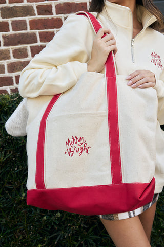 A woman in a light zip-up pullover holds the Merry & Bright Tote Bag—an embroidered canvas tote with red straps and festive design—while standing by a brick wall and greenery.