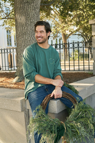 A bearded man with dark hair sits on an outdoor stone ledge, smiling. He wears jeans and a green Winter Sports Gear Long Sleeve Tee, holding a greenery wreath in his lap. Trees and an iron fence are behind him.