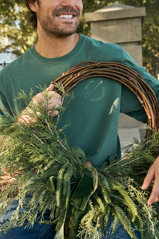A smiling person in a green Winter Sports Gear Long Sleeve Tee holds a pine and twig wreath outdoors on a sunny day, with trees and a stone structure visible in the background.