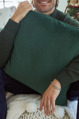 A person in a dark green sweater sits on a white textured blanket, holding the large Repeating Reindeer Pillow in spruce green velvet. Only the lower part of their face is visible; they wear a silver bracelet on one wrist.