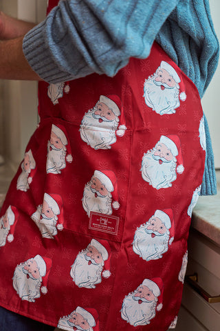 A person in a cozy blue sweater wears the Adult and Child Santa Apron from the Santa Apron + 2 Santa Spatulas Bundle, featuring cartoon Santa faces, while standing in a kitchen with their arm resting on the counter.