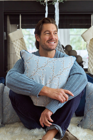 A smiling man with brown hair sits cross-legged on a fluffy rug, hugging the Holly Berry Pillow featuring the exclusive Sewing Down South pattern. He wears a light blue sweater and dark jeans, with stockings and a fireplace in the background.