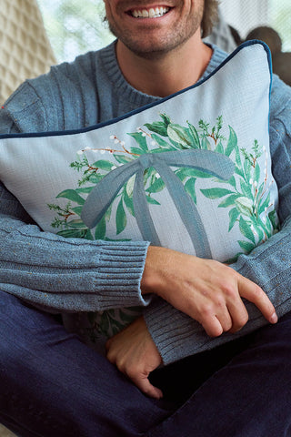 A man in a blue sweater sits cross-legged, holding the Holly Berry Pillow—a festive cushion featuring a green leaf wreath and large letter “T,” exclusively from Sewing Down South. Only his mouth, hand, and part of his body are shown.