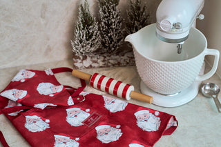 A red apron with Santa Claus prints, a red-and-white striped rolling pin, a stand mixer with a textured white bowl, measuring spoons, and small snowy decorative trees on a kitchen counter.
