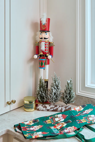 A festive scene with a tall red nutcracker, three small snow-dusted trees, a gold candle, and the Nutcracker Apron featuring an adjustable neck strap, displayed on green nutcracker-print cloth atop a light countertop near a cabinet.