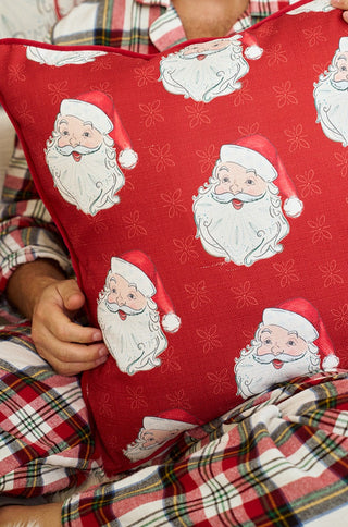 A person in red and white plaid pajamas holds a Vintage Repeating Santa Pillow featuring multiple illustrations of Santa Claus’s face.