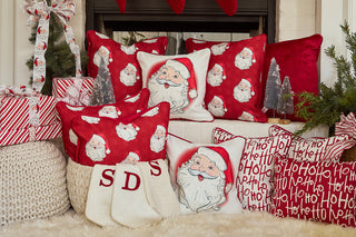 A festive holiday scene with a Vintage Repeating Santa Pillow, red and white pillows featuring Santa faces and “Ho Ho Ho” text, white knit stockings with red initials, wrapped gifts, and small Christmas trees by the fireplace.