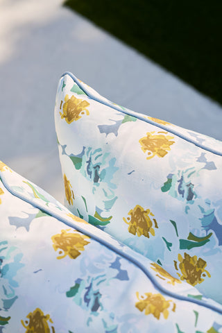 Close-up of two Blossoming Peonies Oversized Outdoor Pillows from Sewing Down South® showcasing floral patterns in blue, green, and yellow. The pillows are artfully arranged on a light surface with grass gently framing the backdrop.