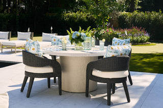 A patio features a round stone table with four wicker chairs that have white cushions and Sewing Down South® floral pillows. A Blossoming Peonies Oversized Outdoor Pillow is on display, alongside decorative items on the table. Sun loungers in the background overlook a lush garden.