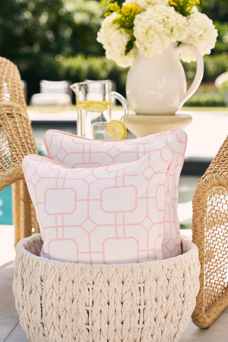 A woven basket holds two pillows, including a Flamingo with Pink Lattice Lumbar Outdoor Pillow, beside a patio pool. A glass pitcher of lemon water and white hydrangeas sit on the table, while wicker chairs and lush greenery provide a serene backdrop.