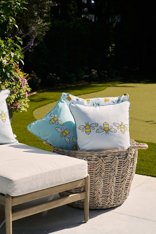 A wicker basket with light blue Bee Trio Lumbar Outdoor Pillows sits on a patio by a cushioned chair. The lush garden backdrop, featuring green grass and diverse plants, enhances this serene setup from our exclusive Outdoor Collection.