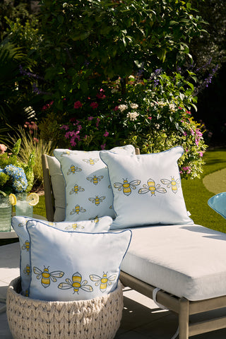 An outdoor lounge area features a beige chaise longue with light blue cushions adorned with Sewing Down Souths yellow and black bee patterns. Nearby, a basket holds matching Bee Trio Outdoor Pillows against lush greenery and colorful flowers in sunlight.