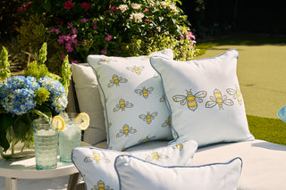 An outdoor patio showcases Sewing Down Souths Bee Trio with Repeating Bees Outdoor Pillow on white cushions. A table nearby holds two glasses of lemon water next to blue hydrangeas. The green lawn in the background highlights durable outdoor fabric that withstands the elements.