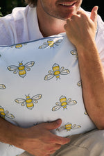 A person holds the Bee Trio with Repeating Bees Outdoor Pillow from Sewing Down South, featuring yellow and white bees on a light backdrop. Crafted with outdoor fabric, it complements their white shirt as they stand outside, face partially visible in the sunlit setting.