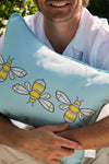 A person in a white shirt, face partially out of frame, holds the Bee Trio Lumbar Outdoor Pillow from the Outdoor Collection. The blue pillow, crafted with SDS Shield® Fabric and featuring three cartoon bees, is set against lush greenery in an outdoor setting.