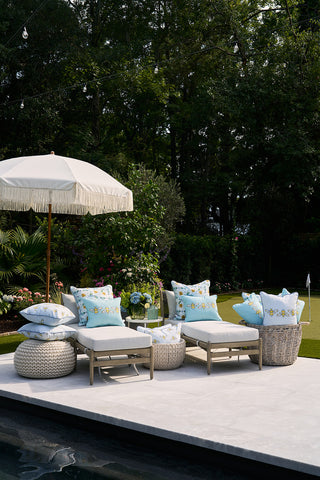 An outdoor patio features light blue cushioned seating and Bee Trio Lumbar Outdoor Pillows, round wicker ottomans by the pool, and a fringed umbrella providing shade. Decorative pillows add comfort while lush greenery surrounds for a serene setting.