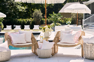 Poolside lounge area with wicker chairs and Flamingo Scatter Outdoor Pillows under umbrellas. A vase of flowers and a glass of lemonade rest on a small table. The sun shines on the nearby pool and greenery, creating a serene outdoor setting.