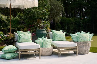Two lounge chairs, each adorned with a Banana Leaves Outdoor Pillow, are positioned next to a basket of extra cushions. The setup includes lush greenery, a large umbrella on the patio, and features Sewing Down South® patterns for added style in a relaxing garden atmosphere.