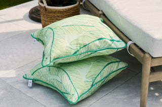 Two Banana Leaves Outdoor Pillows in green leaf patterns are stacked against a light-colored outdoor sofa on a patio. A wicker basket with a plant is visible in the background, creating a relaxed, natural setting with tropical flair.