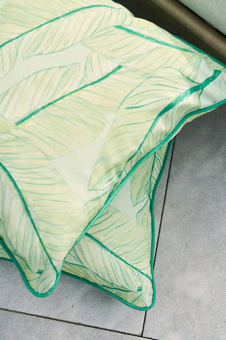 On a gray tiled floor rest two decorative pillows with the Banana Leaves Outdoor Pillow design, showcasing light green leaf patterns and dark green piping. The SDS Shield® Outdoor Fabric provides durability to the pale background, enhancing a light and airy feel.