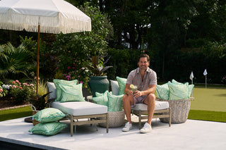 A man sits on a patio chair beneath an umbrella, surrounded by Banana Leaves Outdoor Pillows and potted plants, smiling with an ice cream cone in hand while a putting green is visible behind him.