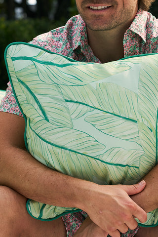 A person in a floral shirt holds the vibrant Banana Leaves Outdoor Pillow, with the Sewing Down South® pattern, while smiling slightly against a blurred green backdrop.