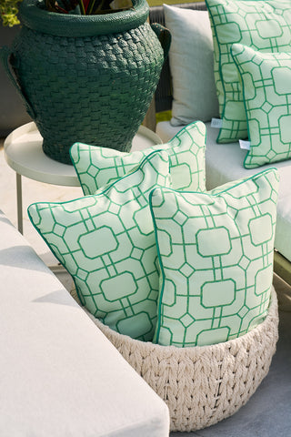 A cozy outdoor seating area is enhanced by a white woven basket holding three Banana Leaves with Green Lattice Lumbar Outdoor Pillows. The scene includes a white cushioned sofa and a textured dark green planter with banana leaves, creating a stylish and comfortable ambiance.