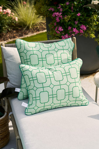 Two Green Lattice Outdoor Pillows, featuring the Sewing Down South® pattern and crafted from SDS Shield® Outdoor Fabric, sit on a light-colored outdoor lounge chair. In the background, large planters host lush greenery and pink blooms.
