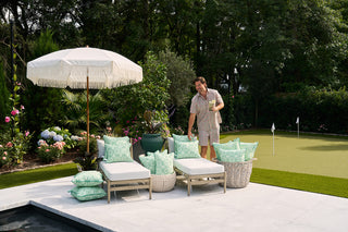 A man arranges Green Lattice Outdoor Pillows made with SDS Shield® Fabric on patio lounge chairs beside a white umbrella, with a putting green and lush greenery in the background.