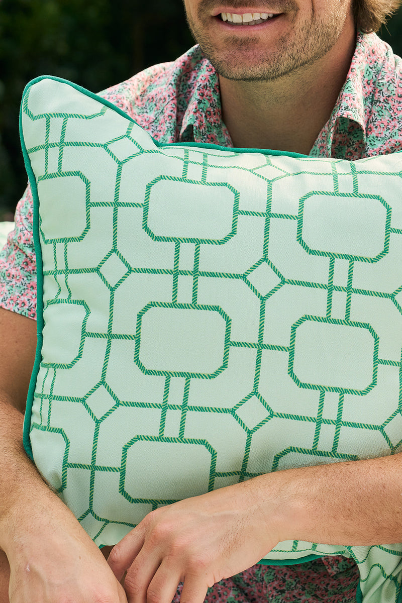 A person wearing a floral shirt holds a Green Lattice Outdoor Pillow from Sewing Down South®, featuring an elegant pattern of interlocking squares in vibrant green on a light background. The focus remains on the stylish pillow rather than their partially visible face.
