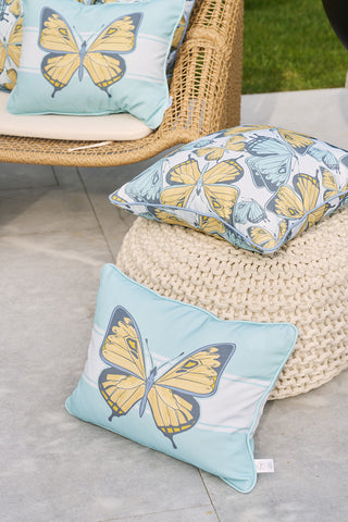 A wicker chair and a knitted pouf display Butterfly Scatter Outdoor Pillows with yellow and blue designs. Theyre set against a stone floor with grass, creating an inviting patio scene.