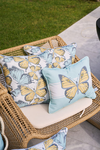 A wicker chair on a gray stone patio is adorned with Sewing Down South® cushions, featuring vibrant yellow and blue butterfly designs against white, and another with a large butterfly on light blue. Perfectly accentuate it using the Butterfly Stripe Lumbar Outdoor Pillow for extra comfort.