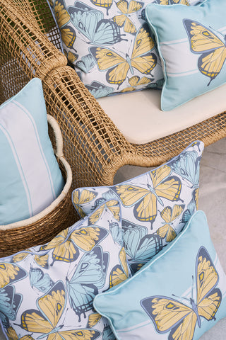 A wicker chair with a white cushion features Sewing Down South® patterns on light blue butterfly scatter outdoor pillows, creating a summery and relaxed setting.