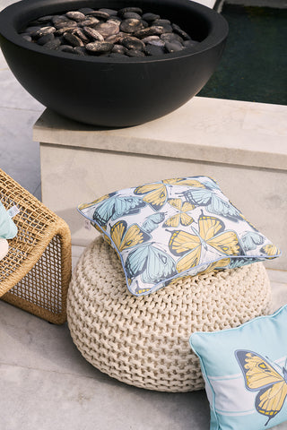 A Butterfly Scatter Outdoor Pillow decorates a knitted pouf next to a wicker chair. A large black bowl of stones rests on the stone patio, enhancing the serene outdoor setting with its Sewing Down South® pattern charm.