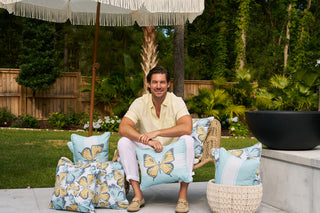 A person in a yellow shirt and white pants relaxes on a patio chair surrounded by Sewing Down South® Pattern pillows, under a fringed umbrella. Greenery and a wooden fence fill the background as they enjoy the comfort of their Butterfly Stripe Lumbar Outdoor Pillow.