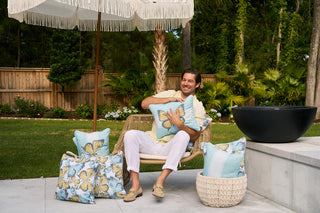 A person sits in a wicker chair outdoors, embracing a Sewing Down South® Butterfly Stripe Lumbar Outdoor Pillow with butterfly designs. Surrounded by more of these pillows and a fringed parasol, the wooden fence and lush greenery enhance the gardens serenity.