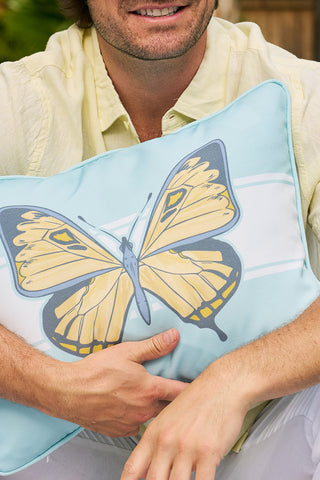 A person in a yellow shirt holds a Butterfly Stripe Lumbar Outdoor Pillow with a delicate yellow butterfly illustration. The persons face is partially visible as they sit outdoors, enjoying the day with their cozy accessory.