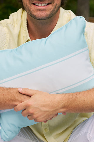 A person in a pale yellow shirt holds a light blue Butterfly Stripe Lumbar Outdoor Pillow with white stripes, smiling warmly from the nose down.