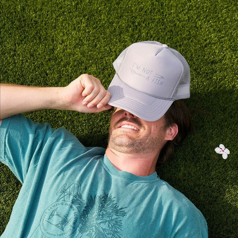 A man is lounging on the grass, his face partly shaded by an Im Not A Fish adjustable snapback hat. The trucker cap pairs with his teal shirt featuring a printed design. A small white flower with a pink center is nearby, adding a whimsical touch of nature.