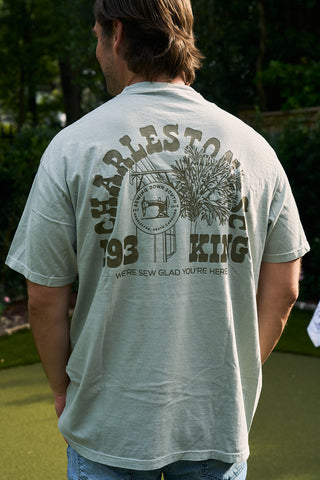A person stands outdoors wearing a light gray SDS 'King Street' Flagship Tee, featuring a large CHARLESTON 193 KING graphic, palm trees, and the text "WERE SEW GLAD YOURE HERE" on the back.