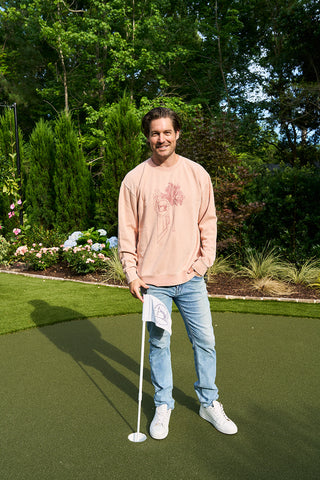 A smiling man in an SDS King Street Illustration Crewneck, jeans, and white sneakers stands on a putting green holding a golf flag. Lush greenery and flowers fill the background.